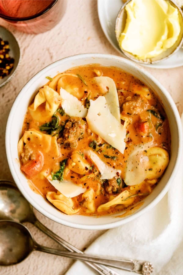 A bowl of creamy tomato pasta soup with tortellini, ground meat, spinach, and slices of parmesan cheese, served with a side of butter.