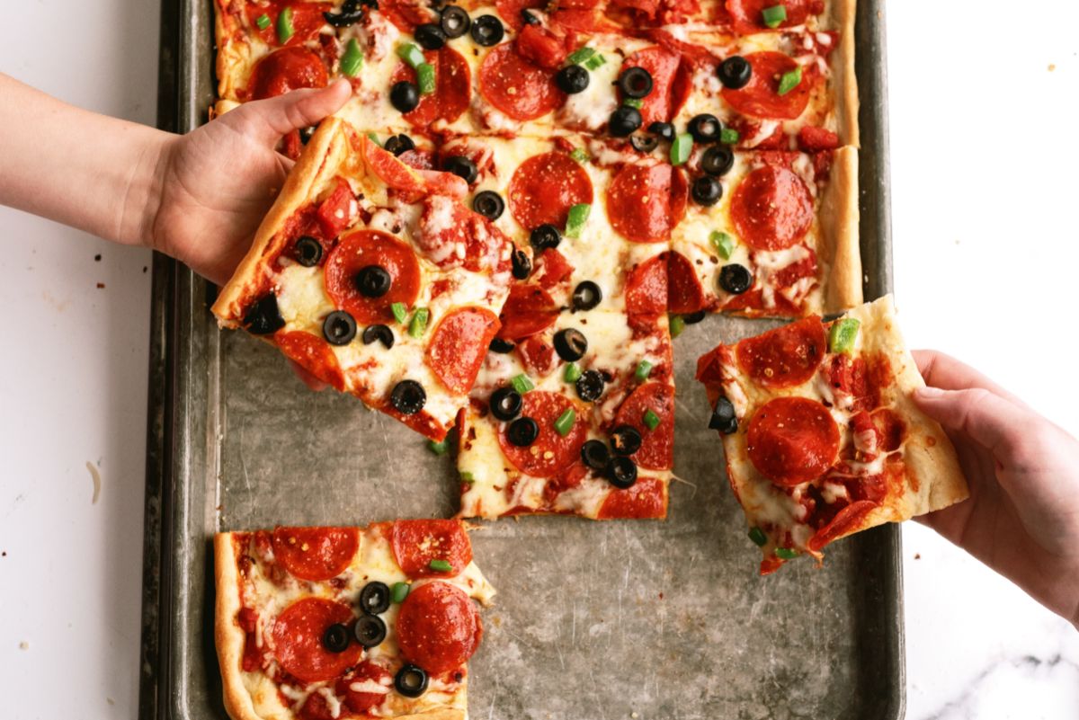 Easy Sheet Pan Pizza cut into squares with 2 people taking pieces