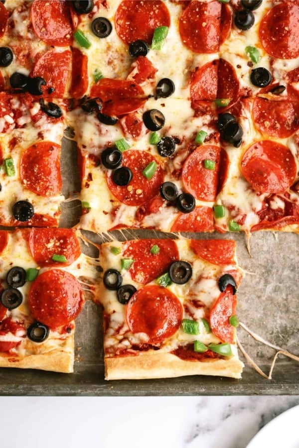 A close-up of a rectangular pepperoni pizza topped with black olives and green bell peppers, divided into square slices.