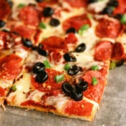 A close-up of a rectangular pepperoni pizza topped with black olives and green bell peppers, divided into square slices.