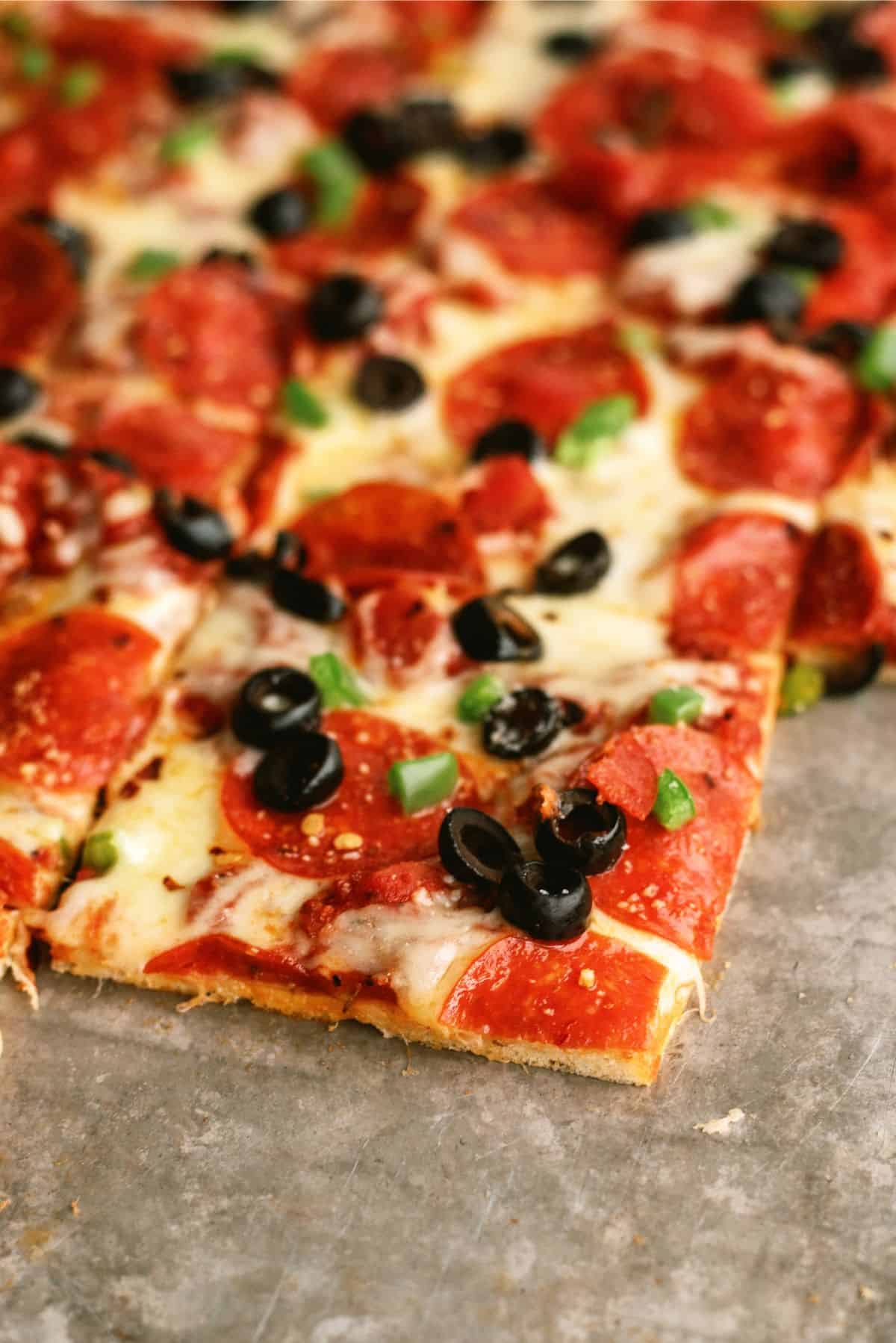 A close-up of a rectangular pepperoni pizza topped with black olives and green bell peppers, divided into square slices.