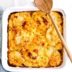 A baked casserole dish with golden, cheesy scalloped potatoes, topped with a wooden spoon resting on the corner.