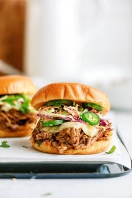 A pulled pork sandwich with coleslaw, sliced jalapeños, and a white sauce in a bun is placed on a white surface.