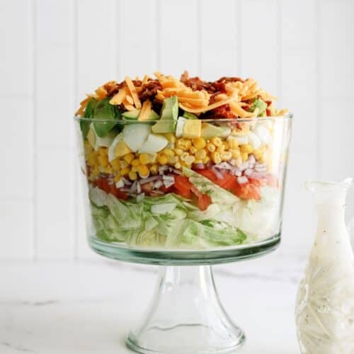 A layered salad in a glass trifle dish with lettuce, tomatoes, onions, corn, cucumbers, cheese, and bacon, next to a glass bottle of dressing.