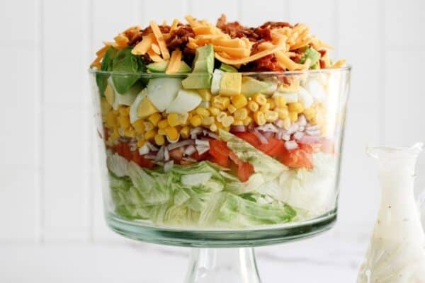 A layered salad in a glass trifle dish with lettuce, tomatoes, onions, corn, cucumbers, cheese, and bacon, next to a glass bottle of dressing.