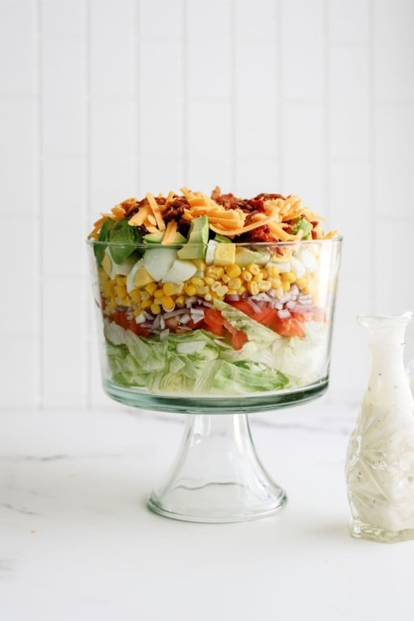 A layered salad in a glass trifle dish with lettuce, tomatoes, onions, corn, cucumbers, cheese, and bacon, next to a glass bottle of dressing.