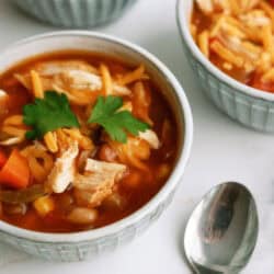 Three bowls of soup containing vegetables, shredded chicken, and garnished with parsley. A metal spoon is placed next to the main bowl.