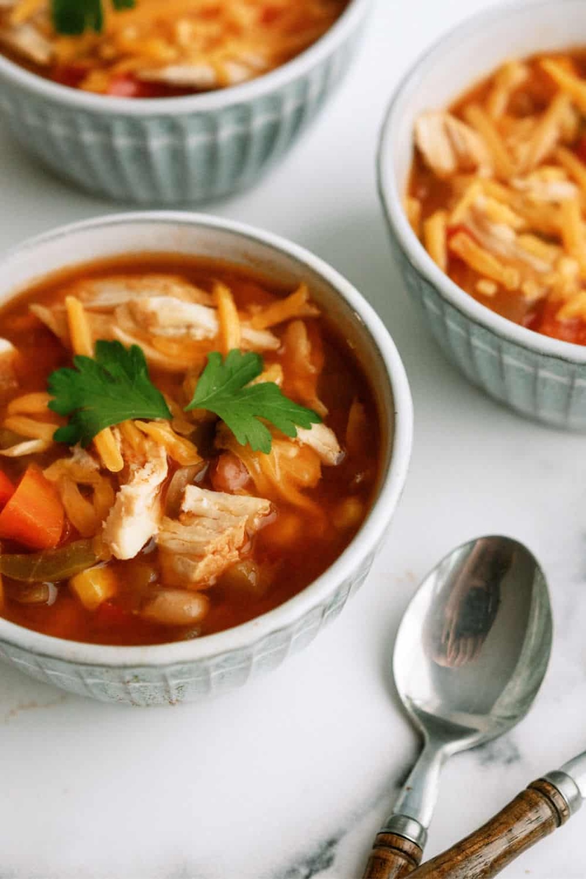 Three bowls of soup containing vegetables, shredded chicken, and garnished with parsley. A metal spoon is placed next to the main bowl.