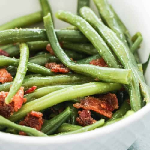 A white bowl filled with cooked green beans and pieces of crispy bacon.