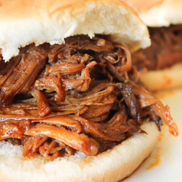 A close-up of a sandwich with shredded barbecue meat filling on a white bun, with another similar sandwich in the background.