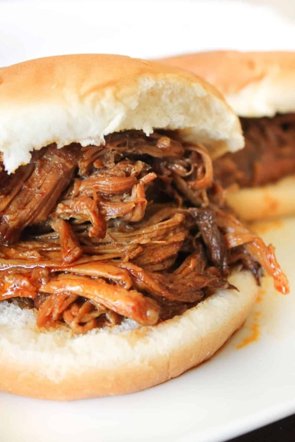 A close-up of a sandwich with shredded barbecue meat filling on a white bun, with another similar sandwich in the background.
