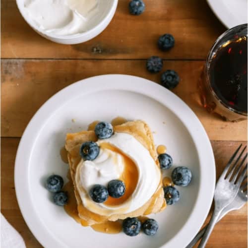 A slice of Baked Pancakes on a white plate topped with berries, whip cream and syrup.