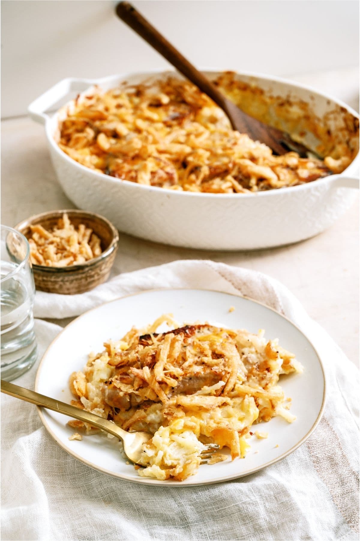 A plate with a serving of Pork Chop Casserole With Hashbrowns on it, and the remainder of the casserole in a dish in the background.