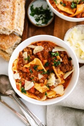 A bowl of hearty lasagna soup garnished with chopped herbs and shaved Parmesan cheese. T