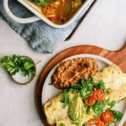 A plate with two enchiladas topped with avocado, cherry tomatoes, cilantro, and a side of refried beans; a casserole dish with more enchiladas is nearby.