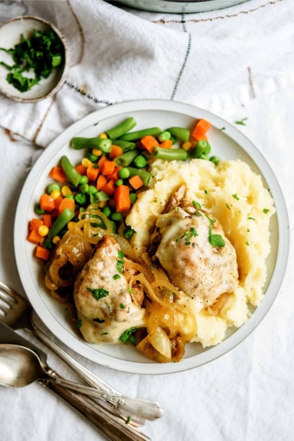 A plate containing mashed potatoes, two pieces of cooked chicken topped with onions, and a side of mixed vegetables including green beans, peas, carrots, and corn.