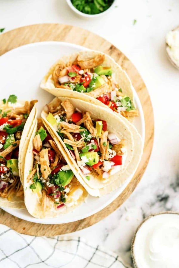 Three tacos filled with shredded chicken, diced tomatoes, onions, cilantro, and cheese served on a white plate on a wooden surface.