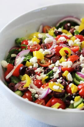A close-up of a salad with sliced red onions, cherry tomatoes, black olives, yellow bell peppers, cucumbers, and crumbled feta cheese in a white bowl.