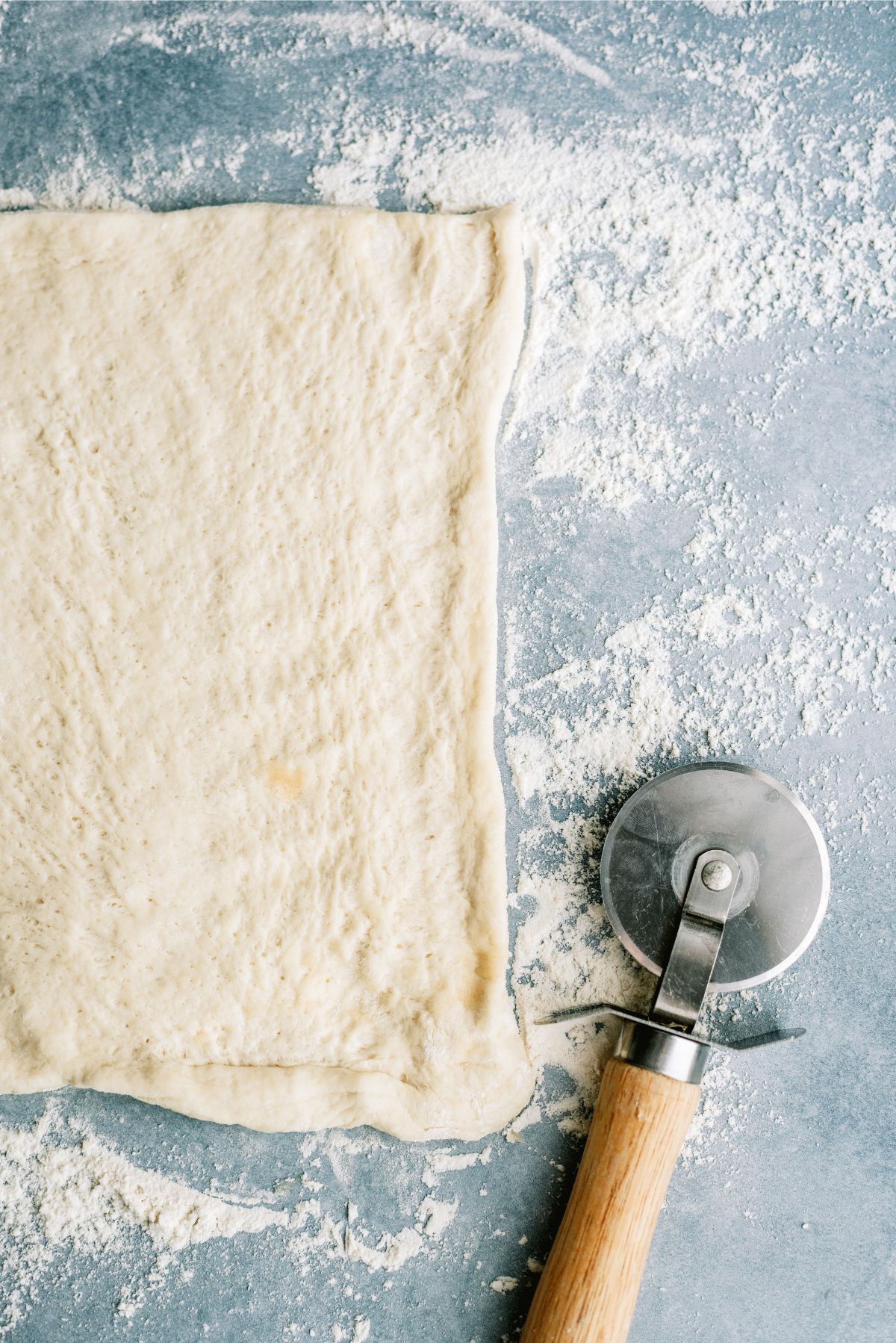 Unrolled pizza dough on a lightly floured surface next to a pizza cutter