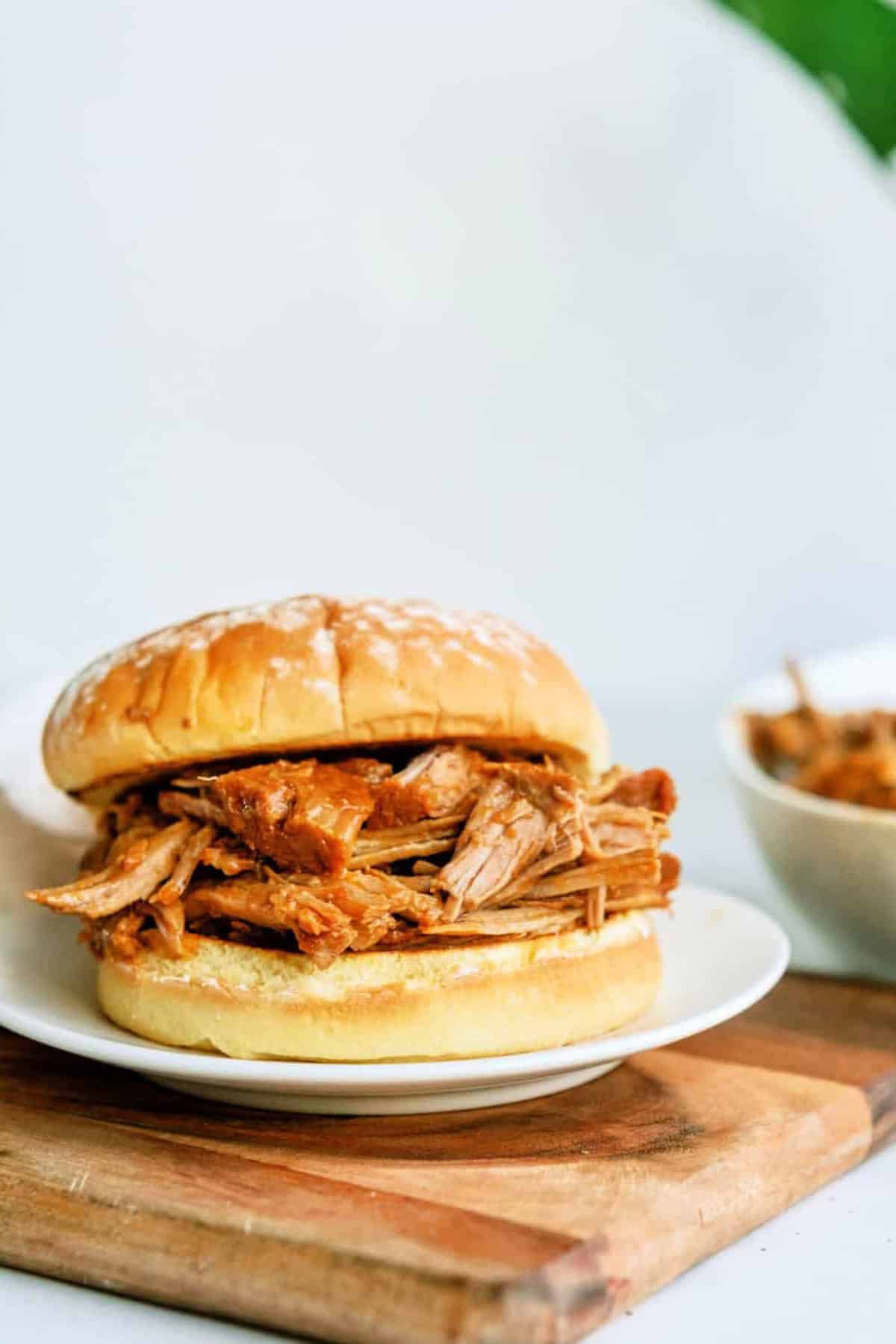 A pulled pork sandwich on a white plate sits on a wooden board, with a bowl of extra pulled pork in the background.