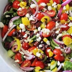 A bowl of Greek salad with cherry tomatoes, cucumbers, red onions, kalamata olives, pepperoncini, and crumbled feta cheese on top.