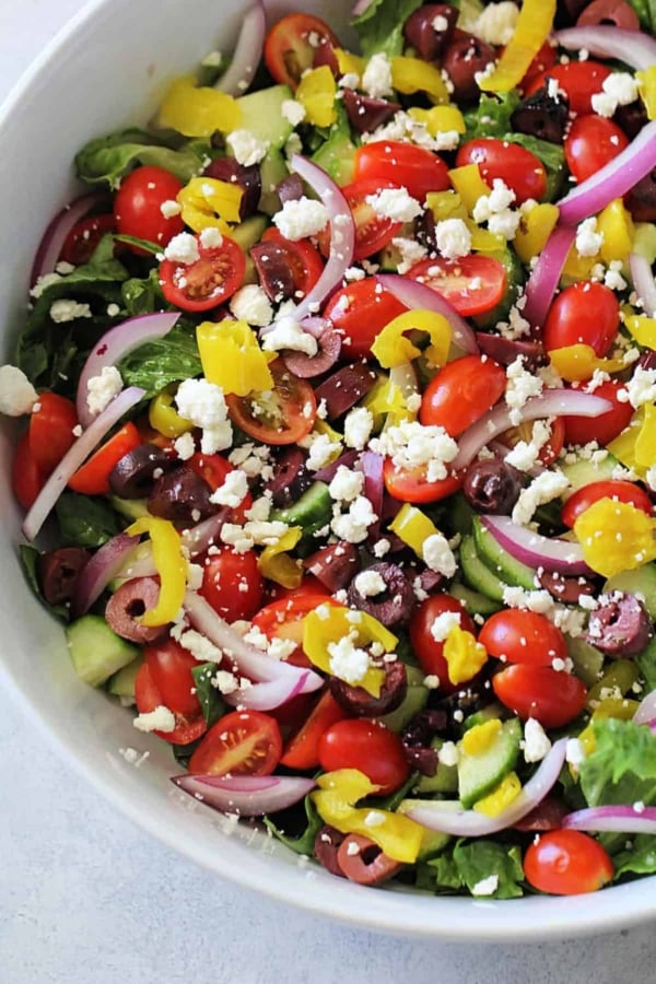 A bowl of Greek salad with cherry tomatoes, cucumbers, red onions, kalamata olives, pepperoncini, and crumbled feta cheese on top.