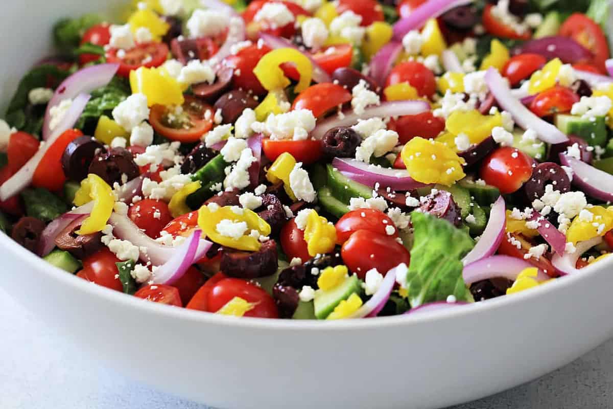 A bowl of Greek salad with lettuce, cherry tomatoes, cucumber, red onion, kalamata olives, pepperoncini, and crumbled feta cheese.