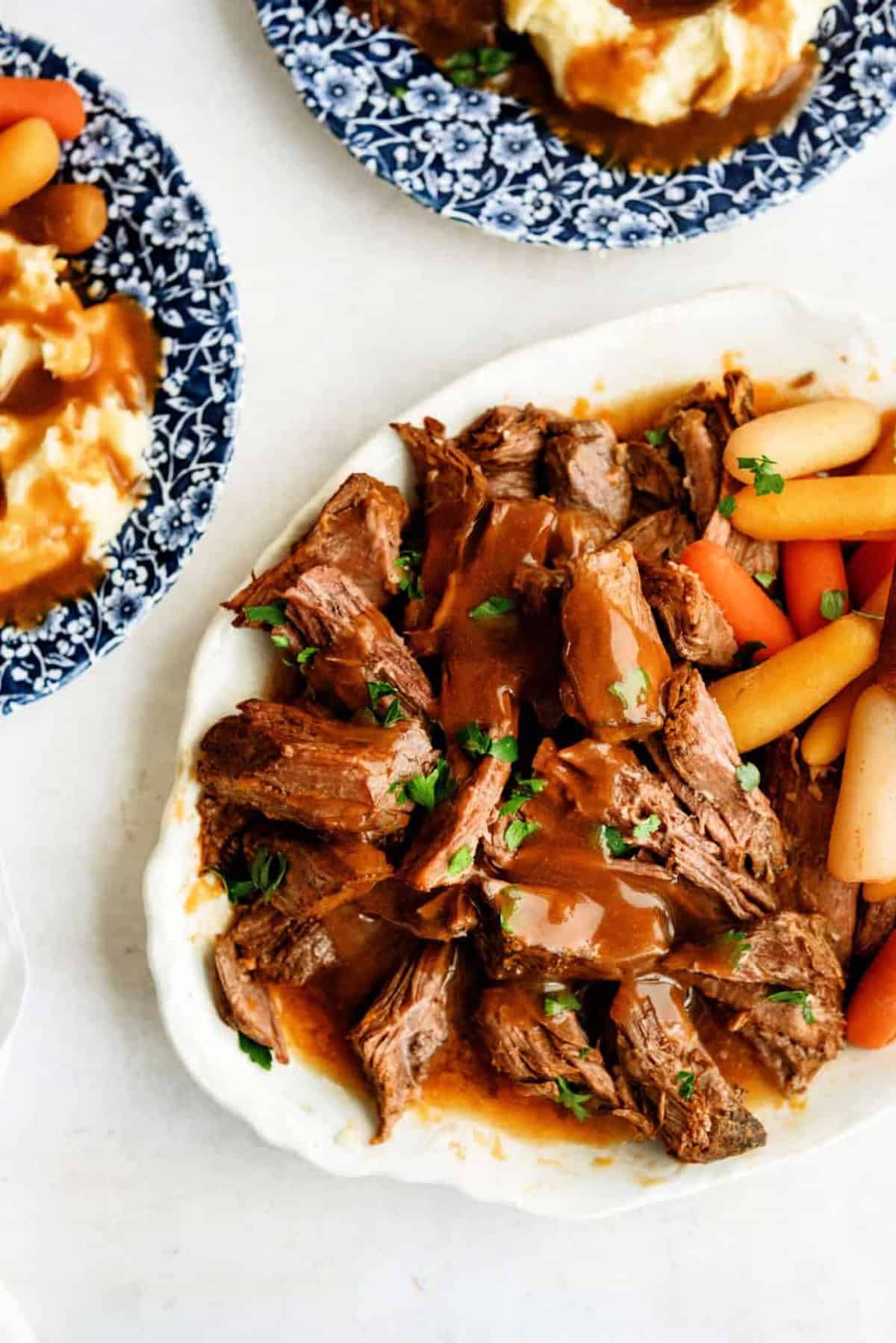 Tender beef pot roast with gravy and vegetables on a white plate.