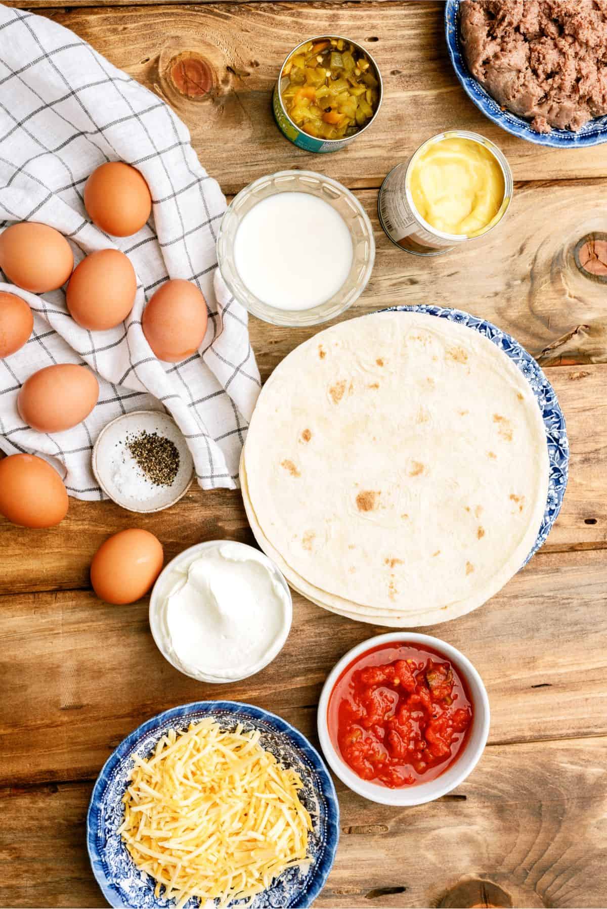 A wooden table with eggs, tortillas, milk, canned goods, ground beef, shredded cheese, salsa, seasoning, and cloth.