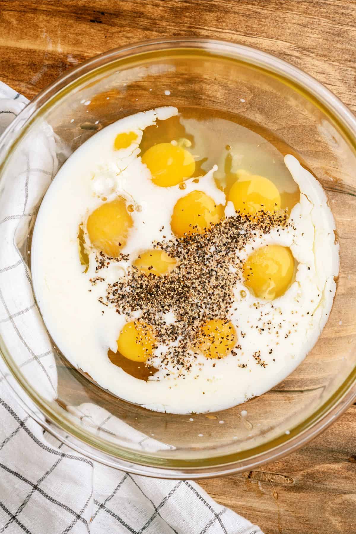 A glass bowl contains cracked eggs, cream, and ground black pepper, placed on a wooden surface next to a checkered cloth.