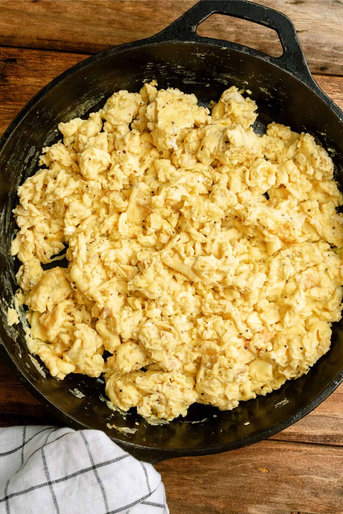 A cast iron skillet filled with scrambled eggs on a wooden surface. A white and black checkered cloth is partially visible at the bottom.
