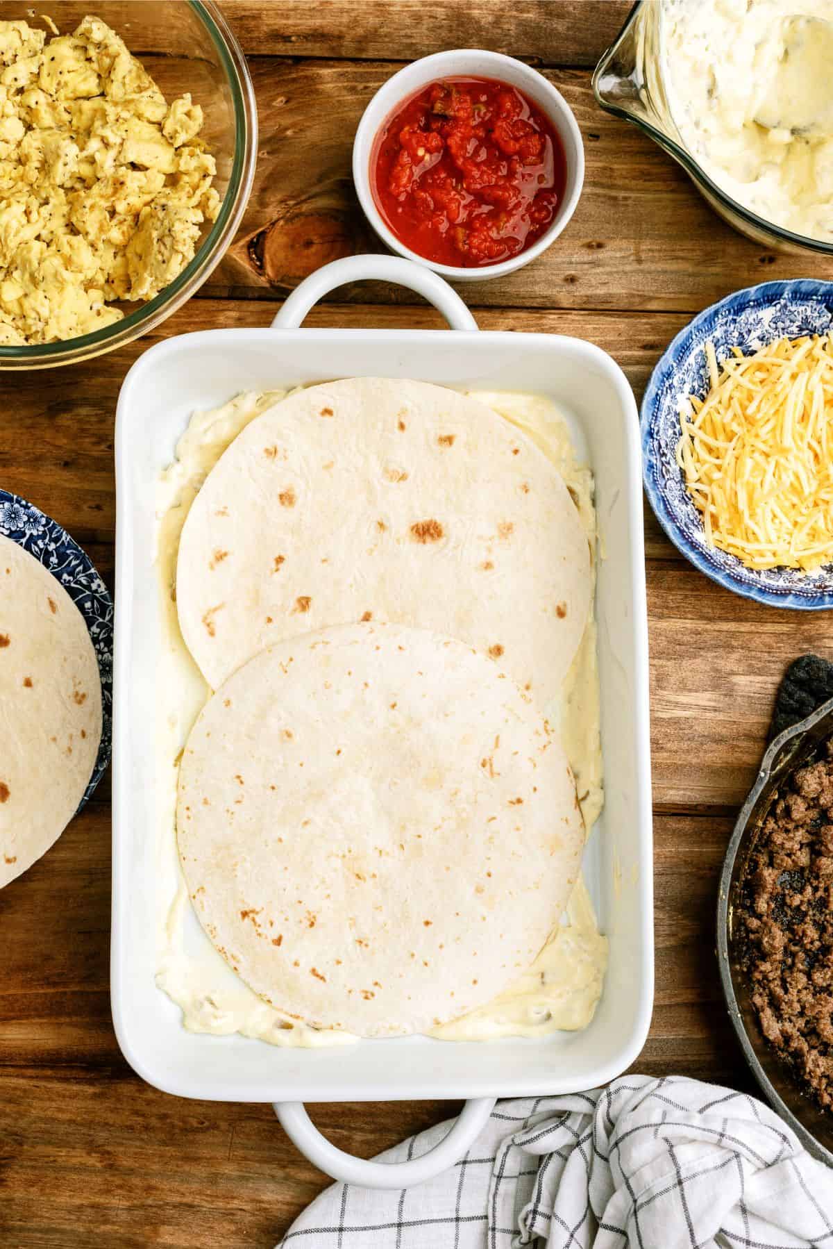 Two flour tortillas are placed in a white baking dish. Surrounding the dish are bowls of scrambled eggs, salsa, shredded cheese, ground meat, and a checked cloth on a wooden surface.