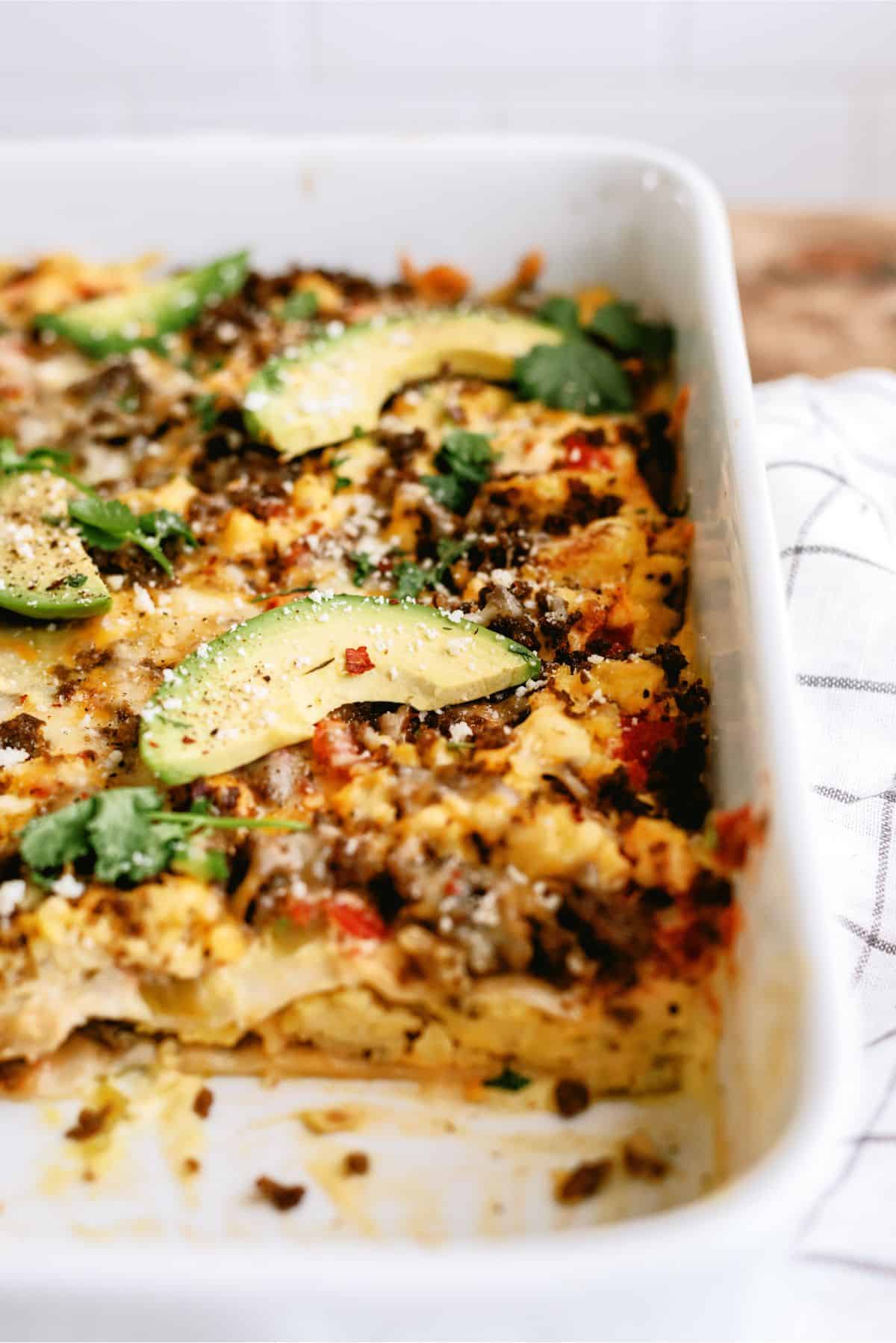 Baked casserole with sliced avocados, herbs, and ground meat in a white dish, with one portion missing.