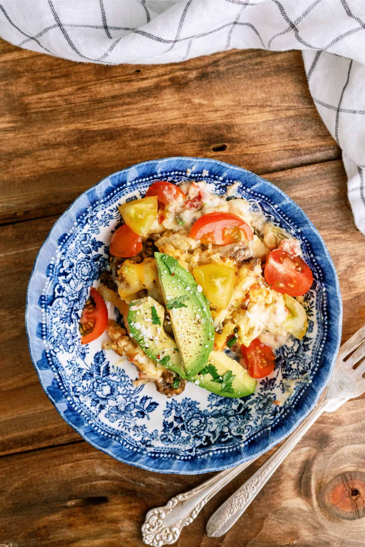 A blue floral-patterned plate with a serving of baked pasta topped with sliced avocado, cherry tomatoes, and herbs. A fork is placed beside the plate on a wooden table with a checkered cloth.