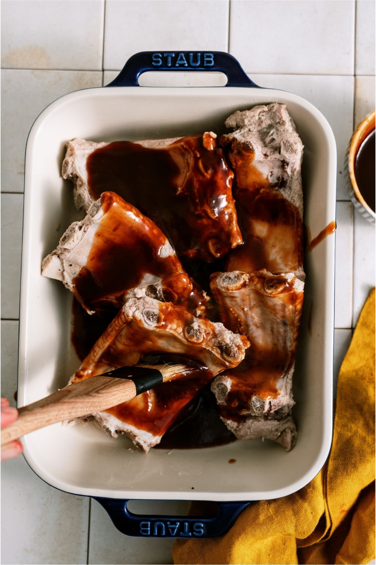 A baking dish filled with rib pieces with a brush, brushing BBQ sauce on top of them.