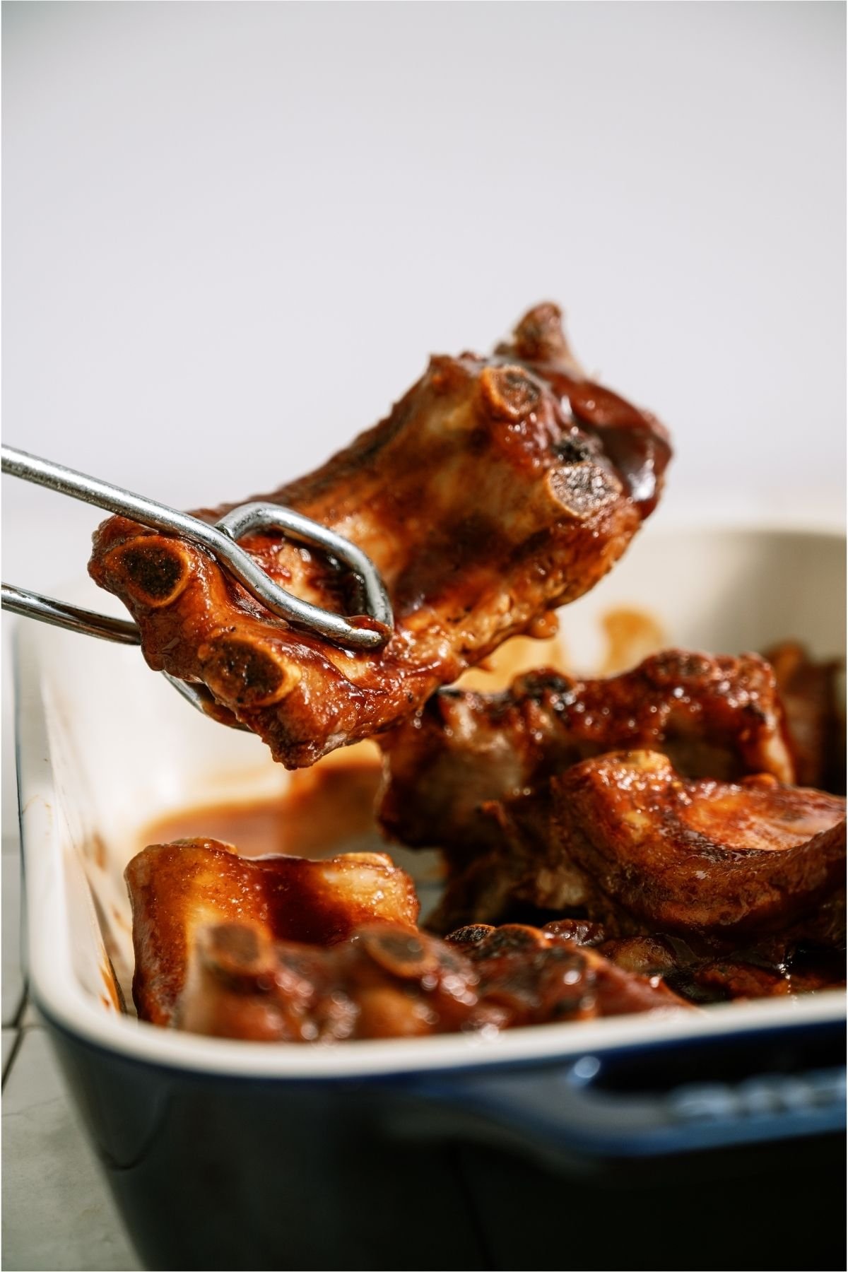 A pair of tongs lifting up a section of Oven Baked BBQ Ribs.