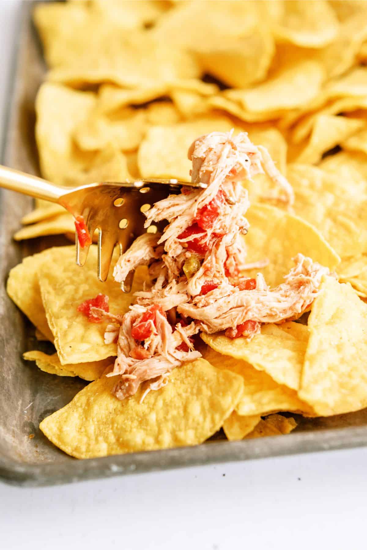 A fork placing shredded chicken on top of tortilla chips in a sheet pan