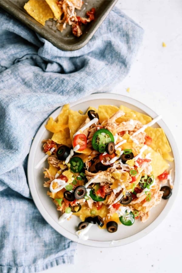 A plate of nachos topped with shredded chicken, sliced jalapeños, black olives, green onions, cherry tomatoes, and drizzled with white sauce, placed on a blue cloth.