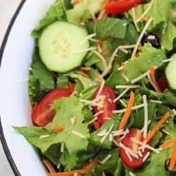 A bowl of salad containing lettuce, cucumber slices, cherry tomato halves, shredded carrots, and grated cheese.