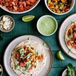 A plate with a taco topped with shredded meat, corn salsa, and green sauce sits on a green tile surface, surrounded by bowls of taco ingredients and lime wedges.