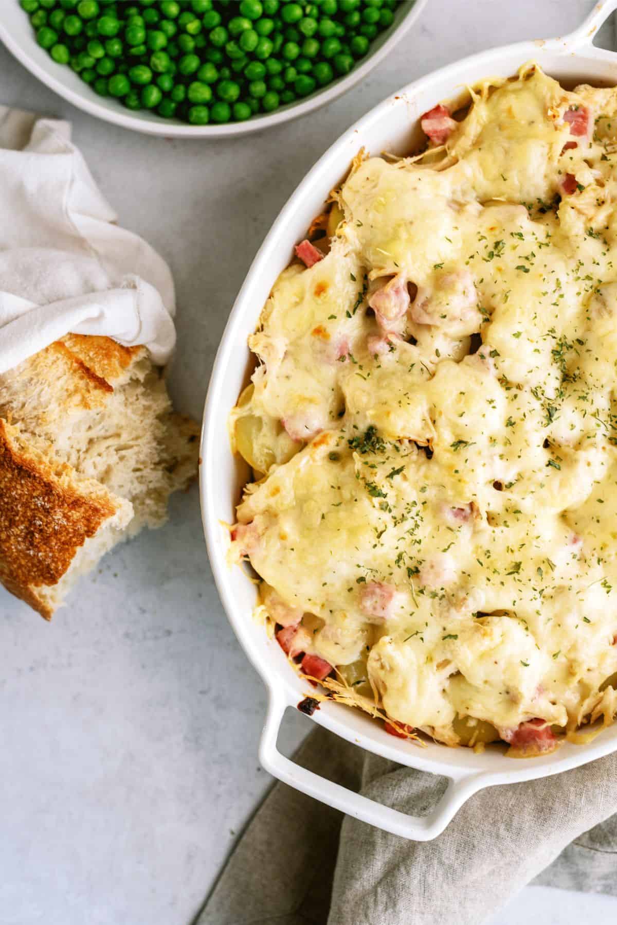 Top view of Chicken Cordon Bleu and Potatoes Casserole in casserole dish with peas and bread on the side