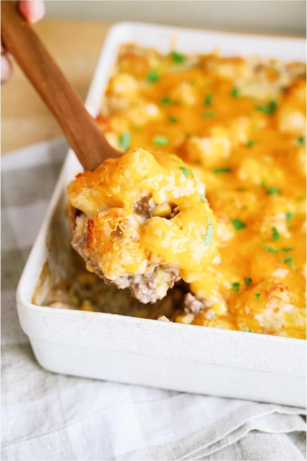 Cowboy Casserole in a large white casserole dish with a wooden spoon lifting out a scoop.