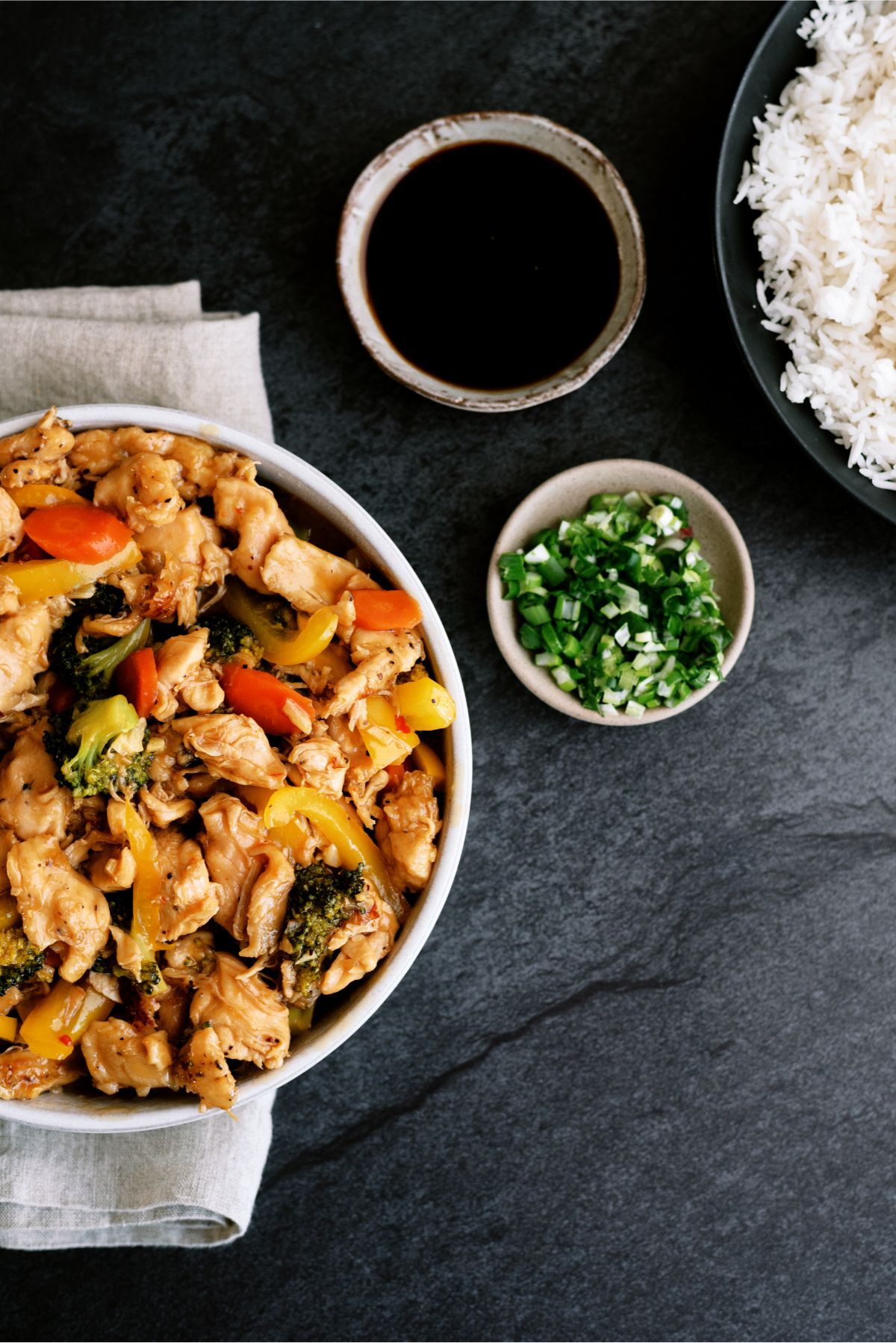 A bowl of Instant Pot Chicken and Veggie Stir Fry, with a bowl of rice, a bowl of sauce and chopped green onions