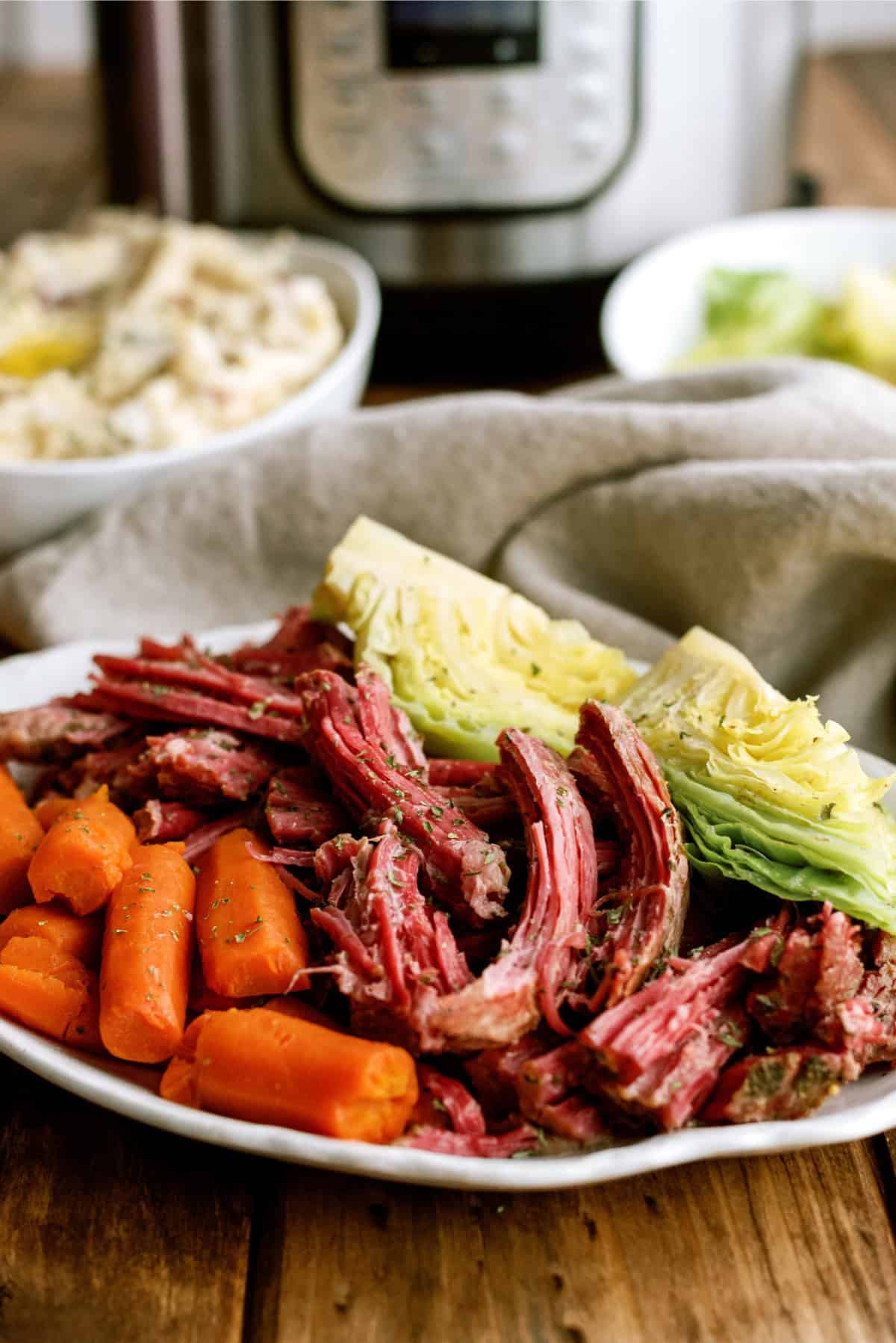 A plate of cooked corned beef, sliced carrots, and wedges of cabbage sits on a wooden table with a bowl of mashed potatoes and an electric pressure cooker in the background.