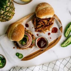 Wooden tray with two pulled pork sandwiches, one topped with red onion and jalapeños, next to a bowl of barbecue sauce, sliced jalapeños, pineapple, and a checkered napkin.