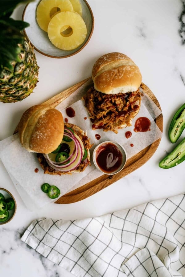 Wooden tray with two pulled pork sandwiches, one topped with red onion and jalapeños, next to a bowl of barbecue sauce, sliced jalapeños, pineapple, and a checkered napkin.