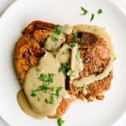 A cooked pork chop topped with creamy gravy and garnished with chopped fresh herbs, served on a white plate.