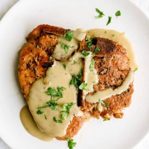 A cooked pork chop topped with creamy gravy and garnished with chopped fresh herbs, served on a white plate.