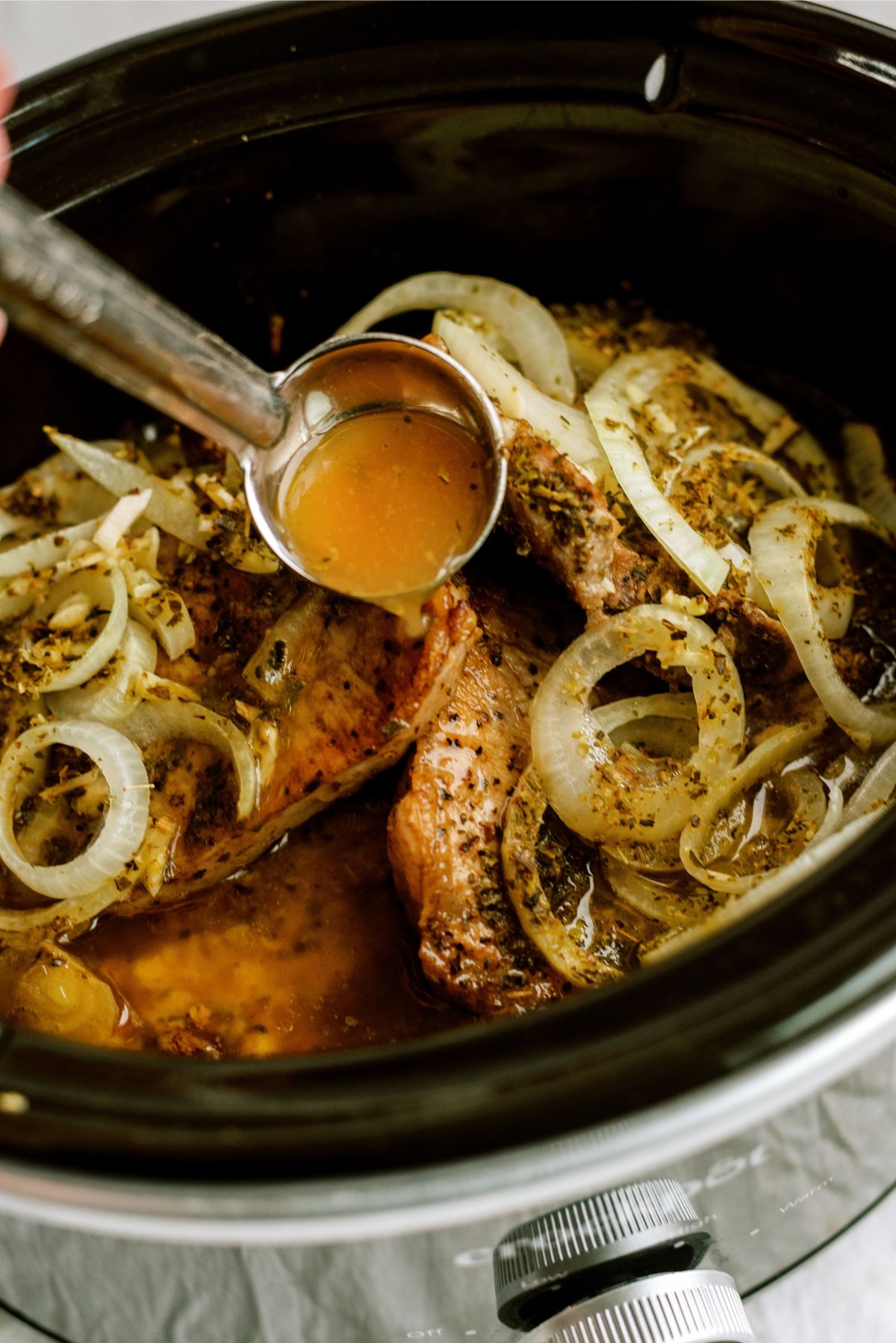 Slow cooker with pork chops and a spoon pouring cooking juices on top.