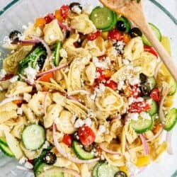 A glass bowl of tortellini pasta salad with cucumbers, cherry tomatoes, artichoke hearts, olives, red onion, and feta cheese, with a wooden spoon.